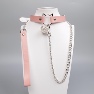 May include: A pink leather choker with a silver cat ear design and a silver chain. The choker has a matching pink leather leash attached. A silver bell charm hangs from the center of the choker.