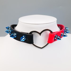 May include: A black and red choker necklace with a silver heart-shaped center. The choker features blue spiked studs. The necklace is displayed on a white mannequin head.