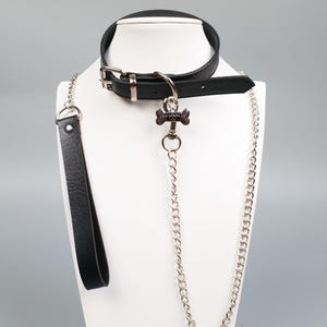 May include: Black leather collar with a silver buckle and a bone-shaped charm. A matching black leather leash and a silver chain are attached. The set is displayed on a white mannequin head.