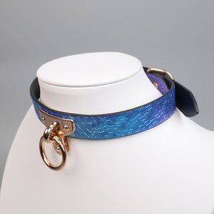 May include: A blue and purple faux snakeskin collar with gold-colored hardware, including a large ring and buckle. The collar is displayed on a white mannequin head, showcasing its design and details.