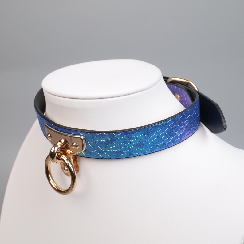 May include: A blue and purple faux snakeskin collar with gold-colored hardware, including a large ring and buckle. The collar is displayed on a white mannequin head, showcasing its design and details.