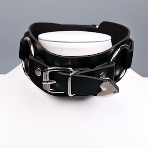 May include: Black leather choker with silver metal hardware. The choker has a heart-shaped accent and an adjustable buckle. The design includes two large metal rings and a double-layered band.