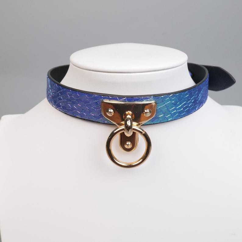 May include: A blue and teal faux snakeskin choker with a gold-colored metal ring and hardware. The choker is displayed on a white mannequin head. The choker has a black backing and a buckle closure.