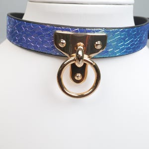 May include: A blue and purple iridescent collar with a gold-colored metal ring and clasp. The collar has a textured, snake-skin-like pattern. The collar is displayed on a white mannequin head.