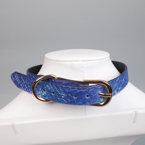 May include: A blue choker-style collar with a textured pattern and a gold-colored buckle. The collar is displayed on a white mannequin head. The collar is made of a blue material with a pattern of white lines.