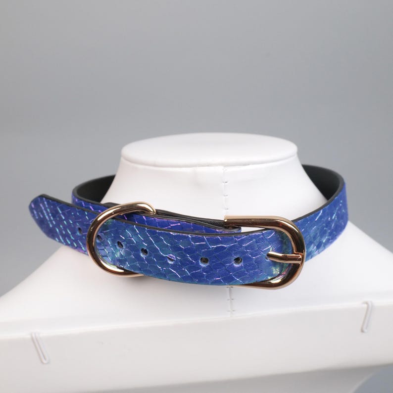 May include: A blue choker-style collar with a textured pattern and a gold-colored buckle. The collar is displayed on a white mannequin head. The collar is made of a blue material with a pattern of white lines.