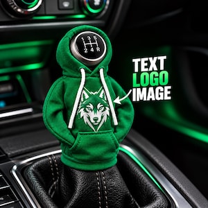 May include: A green miniature hoodie cover for a car gear shift knob, featuring a white wolf head graphic on the front, white drawstrings, and a visible five-speed manual transmission shift pattern on the top of the knob.