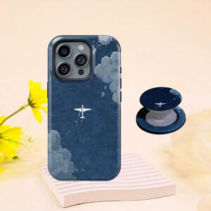 May include: A navy blue phone case and matching pop socket featuring a white airplane design against a cloudy sky. The phone case is shown on a white stand, with a yellow flower in the background.