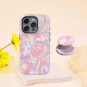 May include: A phone case and matching phone grip with a swirling pink, white, and iridescent design. The phone case is on a white rectangular stand. The phone grip is next to the phone case.