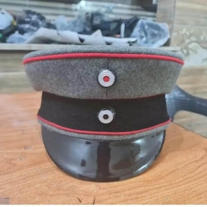 Pode incluir: Um quepe militar cinzento com uma banda preta, pala preta brilhante e debrum vermelho ao longo da coroa e da banda. Duas cocardas circulares com centros brancos, pretos e vermelhos estão posicionadas verticalmente na frente do quepe.
