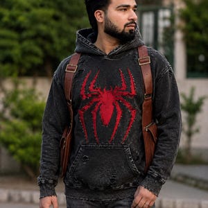 May include: A charcoal black acid-wash hoodie featuring a large red spider graphic on the chest, a front kangaroo pocket, and distressed detailing throughout the fabric, worn with a brown leather harness-style backpack with metallic buckles.