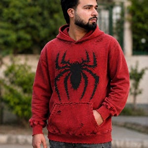 Könnte beinhalten: Ein rot säuregewaschener Kapuzenpullover mit einer großen schwarzen Spinnengrafik auf der Brust. Das Kleidungsstück hat einen Used-Look mit ausgefransten Kanten und kleinen Löchern an Kapuze, Bündchen, Saum und der vorderen Kängurutasche.