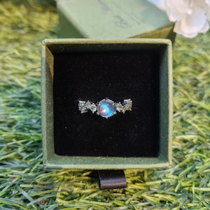 May include: A silver ring with a central, iridescent gemstone, flanked by smaller, clear stones. The ring is displayed in a green velvet box with a black interior, set against a backdrop of green grass.