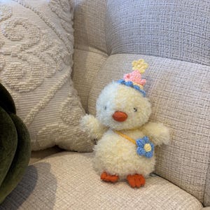 Può includere: Un soffice pulcino di peluche giallo con un cappello da festa con dettagli rosa e blu, un fiore blu e becco e zampe arancioni. Il pulcino è seduto su un divano chiaro.