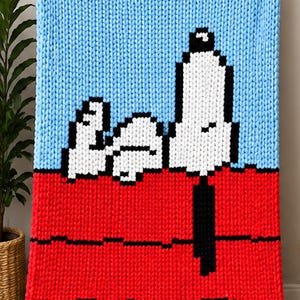 May include: A chunky knit blanket featuring a pixelated graphic of a white dog with black ears and a black nose lying on a red roof against a light blue sky. The blanket is constructed from thick yarn with a visible, textured loop stitch pattern.