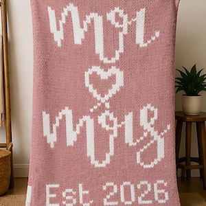 Könnte beinhalten: Eine rosa gestrickte Decke mit den Worten "Mr & Mrs" und einem Herzdesign in Weiß. Die Decke hat auch den Text "Est 2026". Die Decke wird von einer Person gehalten.
