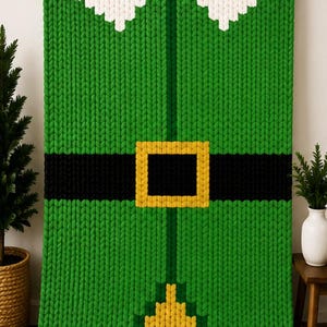 May include: A chunky knit blanket featuring a green base with a white collar design at the top, a horizontal black stripe across the middle, a yellow square buckle detail, and a yellow triangular pattern at the bottom edge, hanging on a rod.