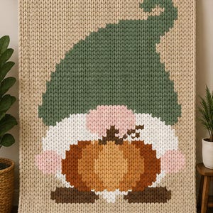May include: A chunky knit blanket in beige featuring a large gnome graphic. The gnome has a tall green hat, a white beard, pink cheeks, and is holding a brown pumpkin. The design is created with thick yarn stitches in a textured, woven pattern.