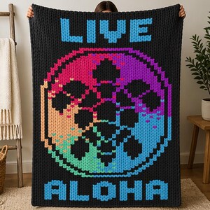 Peut inclure: Couverture noire tricotée avec les mots "LIVE ALOHA" en lettres bleues. La couverture présente un motif pixelisé coloré en forme de cercle avec des teintes arc-en-ciel de rose, orange, vert et bleu.