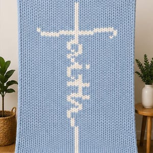 May include: A light blue knitted blanket with the word "Faith" in white, vertically centered, and a white cross at the top. The blanket is made of thick, chunky knit yarn. The blanket is displayed on a light background.