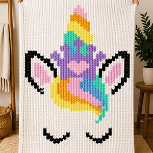 Puede incluir: Una gran manta de punto blanco roto con un diseño de unicornio colorido. El cuerno del unicornio es un arcoíris de colores, y la cara incluye pestañas negras y orejas con detalles en rosa. La manta se sostiene, mostrando su tamaño y el detallado patrón de punto.