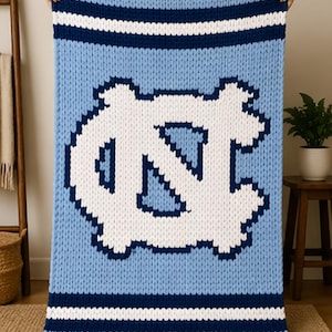 Peut inclure: Une grande couverture en tricot épais bleu clair avec un motif blanc et bleu foncé. La couverture présente un grand logo "NC" blanc au centre, avec des rayures bleu foncé et blanches en haut et en bas. La couverture est tenue par des mains invisibles.