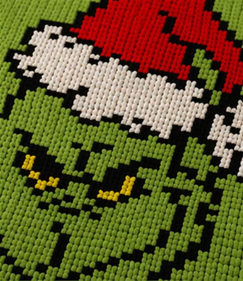 May include: Close-up of a green, red, white, black, and yellow pixelated design. The design features a character's face with a red hat, white face, black eyes, and a green background.