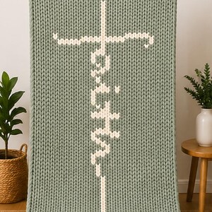May include: A sage green knitted blanket with the word "Faith" in white, vertically centered. The word is designed with a cross at the top. The blanket is displayed indoors.
