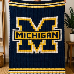 May include: A navy blue knitted blanket featuring the word "MICHIGAN" in a white and yellow block letter design. The blanket has yellow and white stripes at the top and bottom. The blanket is held up by a person.
