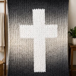 May include: A large, chunky-knit blanket with a gradient design, transitioning from black at the bottom to white in the center, forming a prominent cross shape. The blanket is displayed on a wall, showcasing its texture and design.