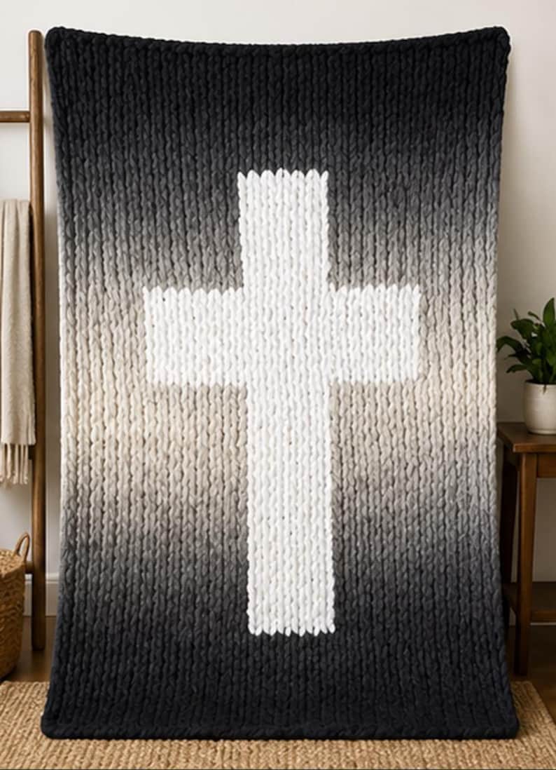 May include: A large, chunky-knit blanket with a gradient design, transitioning from black at the bottom to white in the center, forming a prominent cross shape. The blanket is displayed on a wall, showcasing its texture and design.