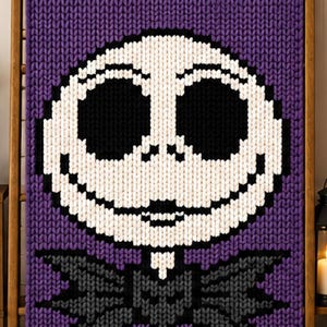 May include: A chunky knit blanket featuring a large, central white skull face with oversized black eye sockets, a small nose, and a wide stitched smile. Below the head is a dark gray bat-shaped bowtie, all set against a solid purple background.