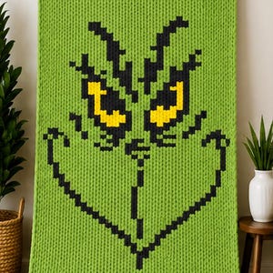 May include: A lime green chunky knit blanket featuring a pixelated graphic of a character face. The design is rendered in black yarn for the eyebrows, eyes, nose, and mouth, with bright yellow yarn filling the center of the two eyes.