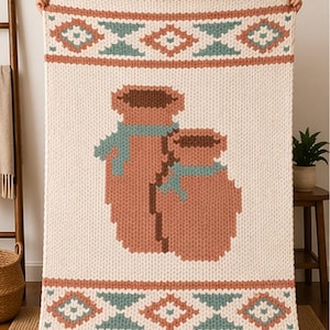 May include: Cream-colored knitted blanket with a geometric border in rust and teal. The center features two brown vases with teal scarves. The blanket is displayed against a neutral background, showcasing its cozy texture and design.