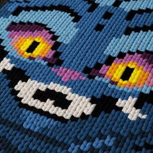 May include: Close-up of a blue, pixelated cat face with a wide grin. The cat's eyes are yellow with purple and pink accents. The design is made of small, textured squares, creating a 3D effect. The cat's fur is various shades of blue.