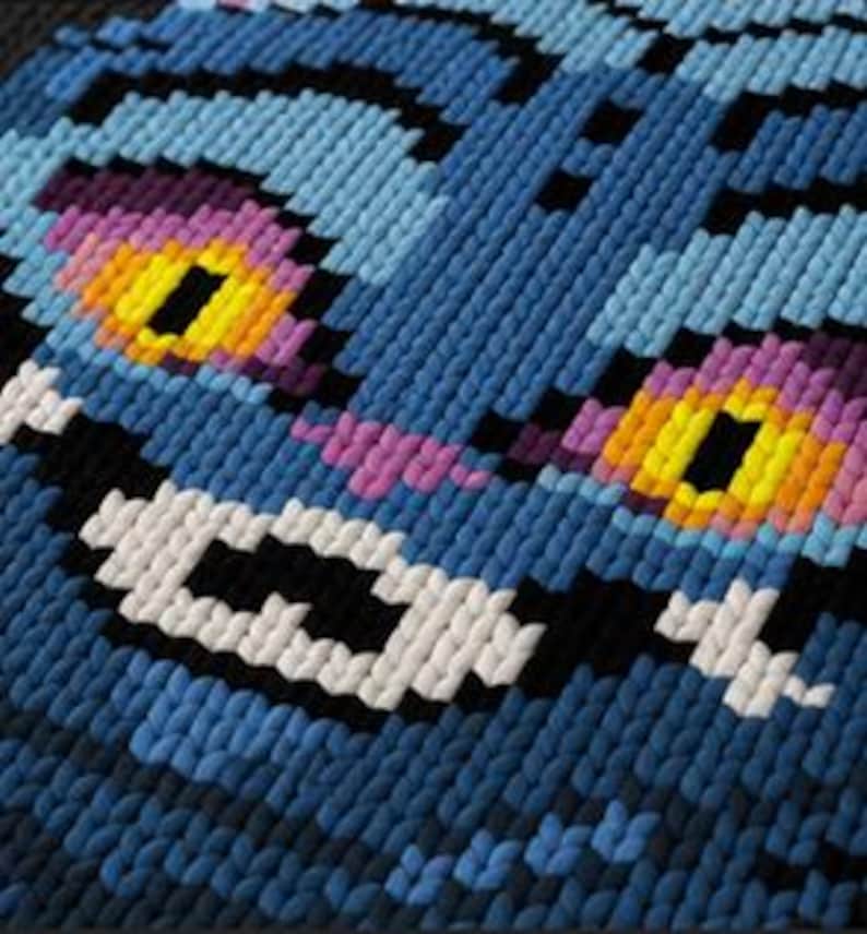 May include: Close-up of a blue, pixelated cat face with a wide grin. The cat's eyes are yellow with purple and pink accents. The design is made of small, textured squares, creating a 3D effect. The cat's fur is various shades of blue.