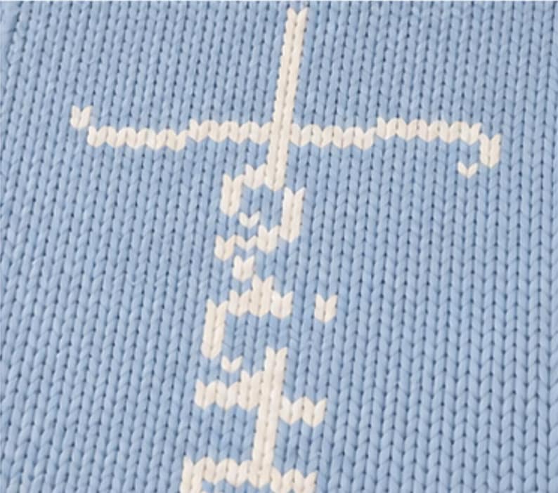 May include: Close-up of a light blue knitted fabric with a white cross design. The cross is made of a series of small, connected stitches, creating a textured appearance. The fabric has a visible knit pattern.