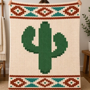 May include: Cream-colored knitted blanket featuring a large green cactus design. The blanket has a geometric border in shades of brown and teal. The blanket is held up by a person.