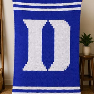 Pode incluir: Um cobertor de malha azul e branco com uma grande letra "D" branca no centro. O cobertor tem duas listras brancas na parte superior e inferior. O cobertor é segurado por uma pessoa.