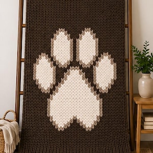 May include: A brown knitted blanket with a large paw print design in cream and light brown. The blanket is displayed on a wooden ladder, showcasing its texture and pattern. The cozy, pet-themed blanket is a decorative home accessory.