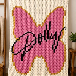 May include: A chunky knit blanket in cream yarn featuring a large pink butterfly shape outlined in mustard yellow. The center of the butterfly displays the word "Barbie" written in black cursive script, integrated into the knitted texture of the fabric.
