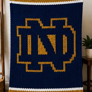 May include: A navy blue knitted blanket featuring a gold and white striped border and a large gold "ND" logo in the center. The blanket is made of thick yarn and is held up by a person.