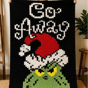 Op de afbeelding: Zwarte gebreide deken met de woorden "Go Away" in het wit, een rode kerstmuts en een groen gezicht met gele ogen. De deken wordt door een persoon omhoog gehouden.