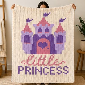 May include: Cream-colored knitted blanket featuring a purple and pink castle design with flags. The blanket has the words "little princess" in pink and purple. The blanket is held up by a person.