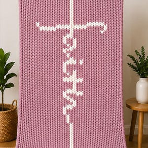 May include: A chunky knit blanket in a dusty rose color. The blanket features a white cross design with the word "Faith" vertically stitched in the center. The blanket is displayed on a neutral background.