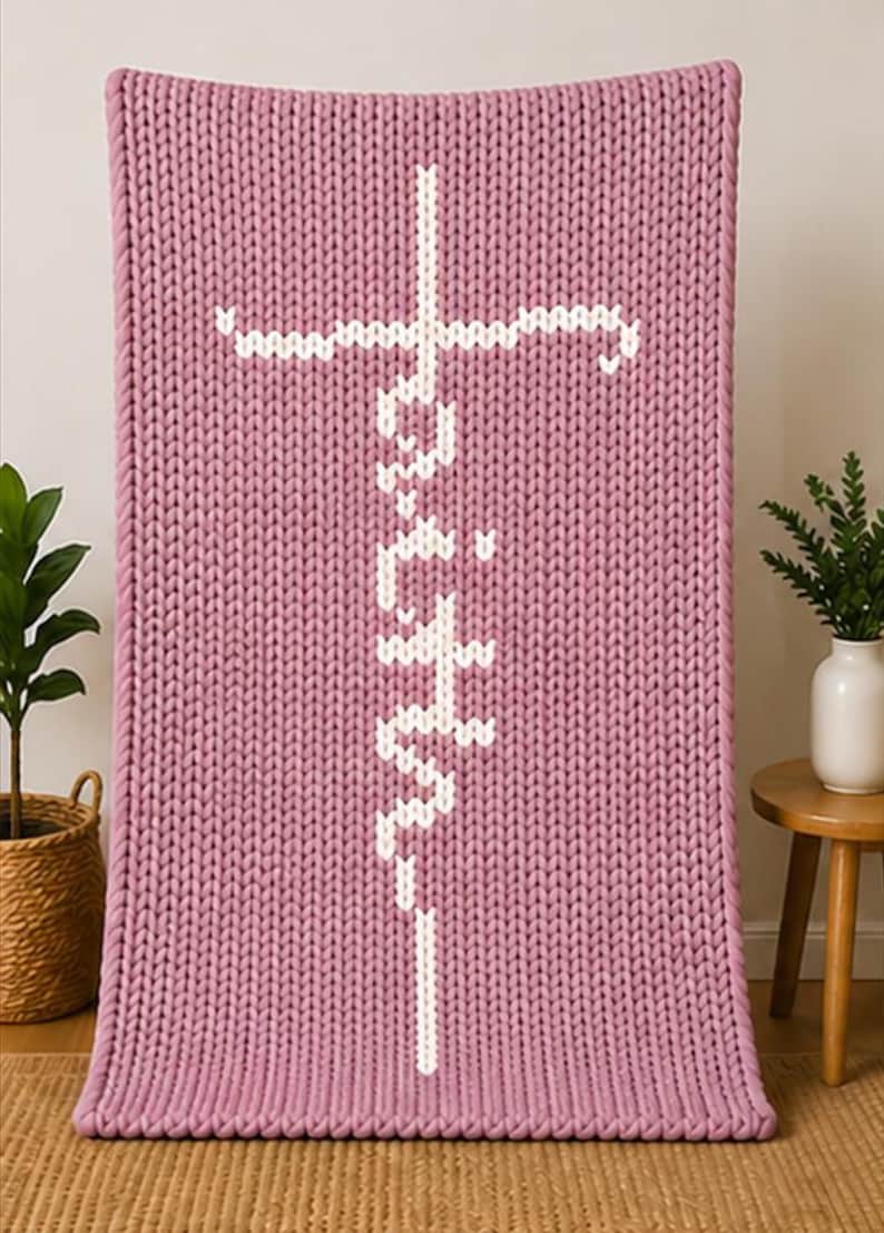 May include: A chunky knit blanket in a dusty rose color. The blanket features a white cross design with the word "Faith" vertically stitched in the center. The blanket is displayed on a neutral background.