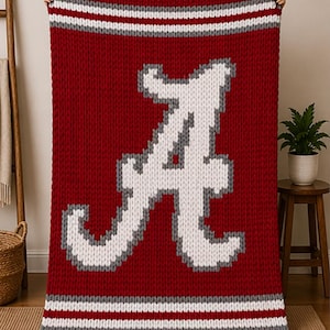 May include: A large, knitted red blanket with a white and gray letter "A" in the center. The blanket has white and gray stripes at the top and bottom. The blanket is made of thick yarn.