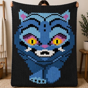 Puede incluir: Una gran manta de punto negra con la cara de un gato azul con ojos amarillos, orejas rosas y una amplia sonrisa. El diseño del gato está pixelado, lo que le da una apariencia texturizada única. La manta está sujeta por una persona.