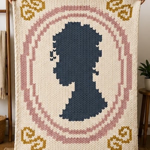 Puede incluir: Manta de punto color crema con una silueta azul dentro de un marco ovalado rosa y crema. La manta tiene elementos decorativos dorados en cada esquina. El diseño recuerda a un retrato vintage.