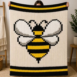 May include: Cream-colored knitted blanket featuring a large, pixelated bee design in black and yellow. The blanket has black and yellow stripes at the top and bottom. The bee has white and gray wings.